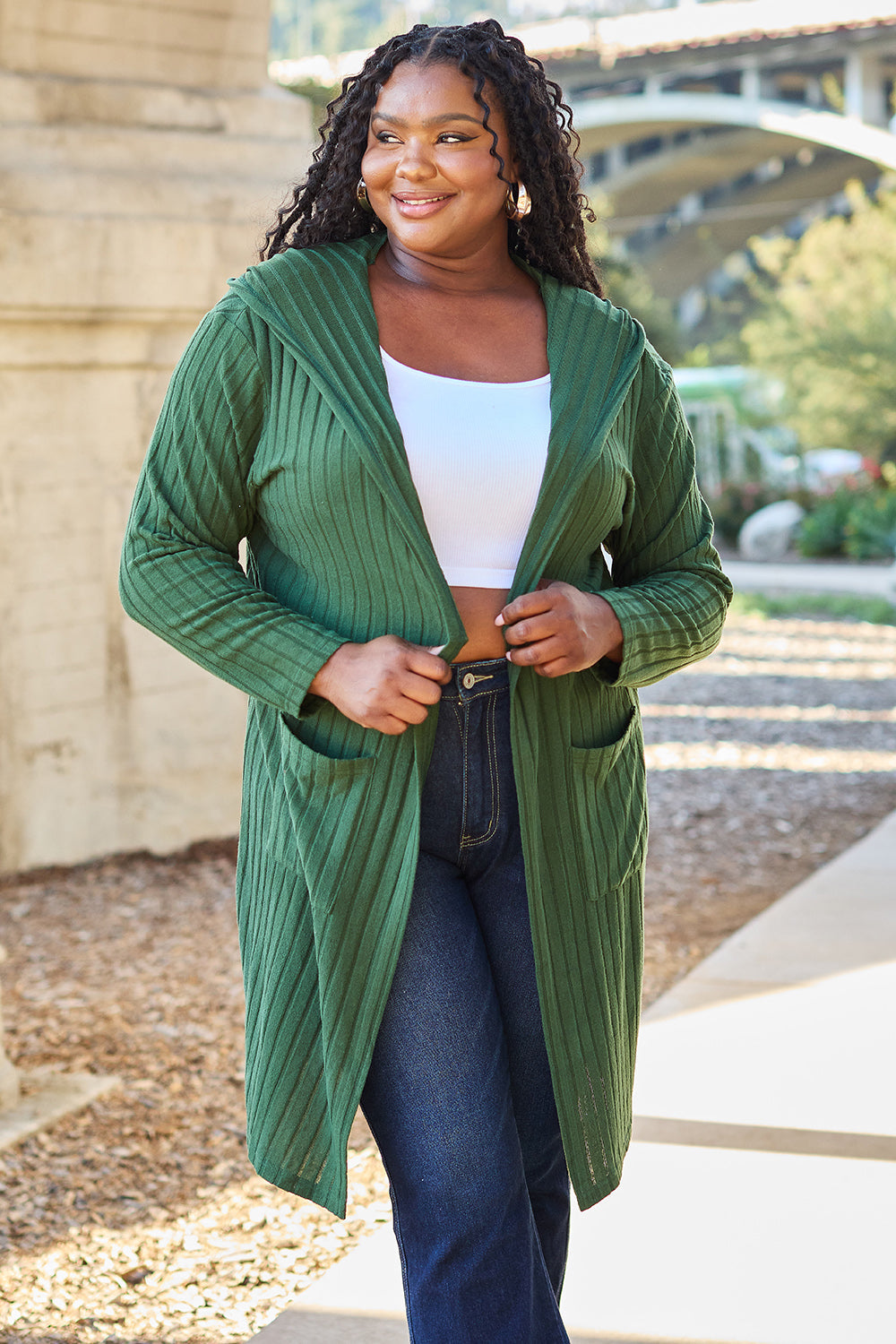 Woman wearing a green hooded sweater cardigan with pockets, paired with jeans and a white top, enjoying a sunny outdoor setting.