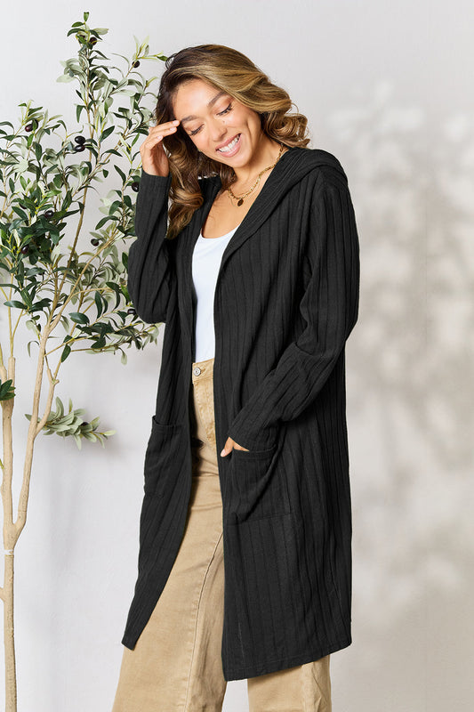 Woman wearing a black hooded sweater cardigan with pockets, paired with a white top and beige pants, standing by a leafy plant.