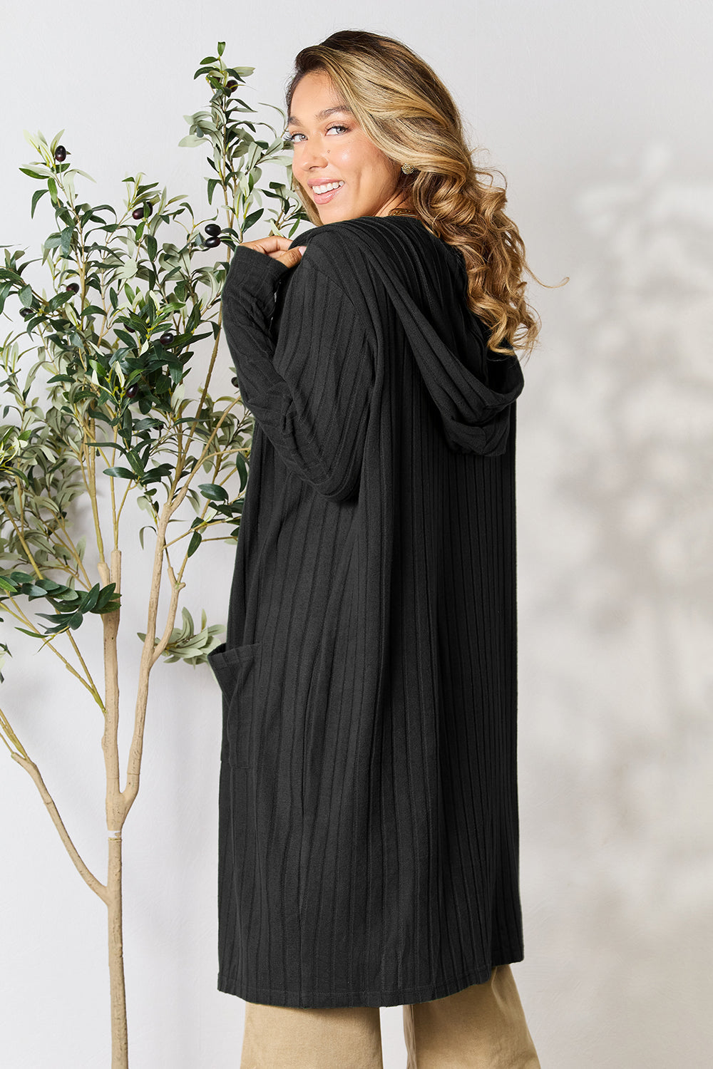 Woman wearing a black hooded sweater cardigan with pockets, standing by a plant, showcasing the back design and slight stretch of the fabric.