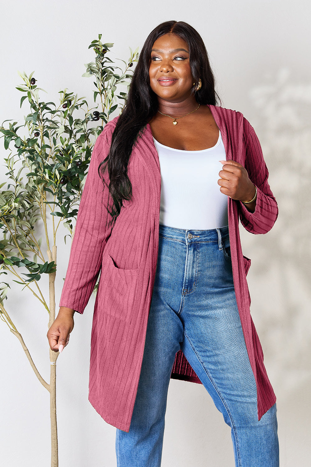 Woman wearing a pocketed hooded sweater cardigan in pink with a white top and jeans, standing next to a plant.