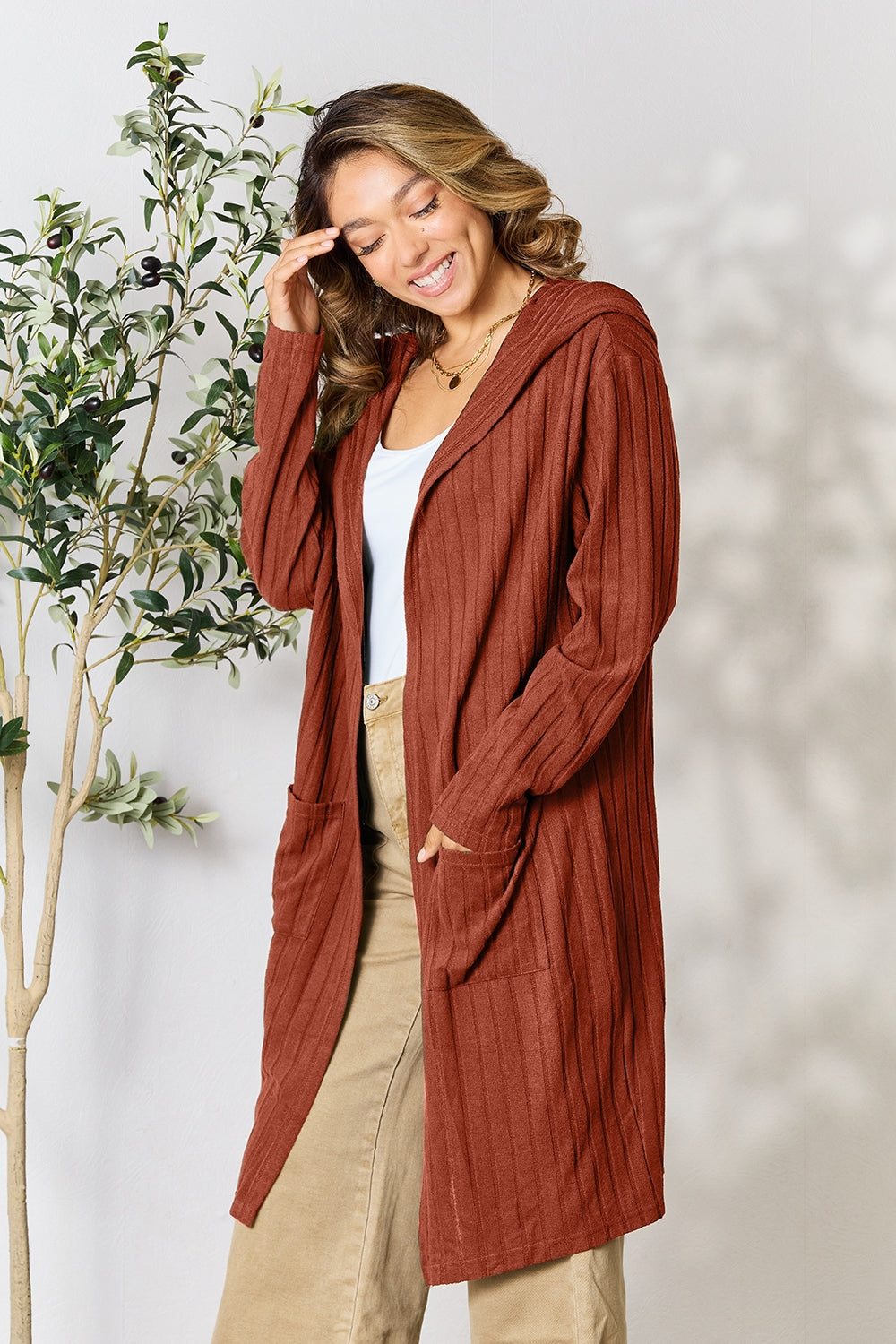 Woman wearing a rust-colored hooded sweater cardigan with front pockets, styled with a white top and beige pants.