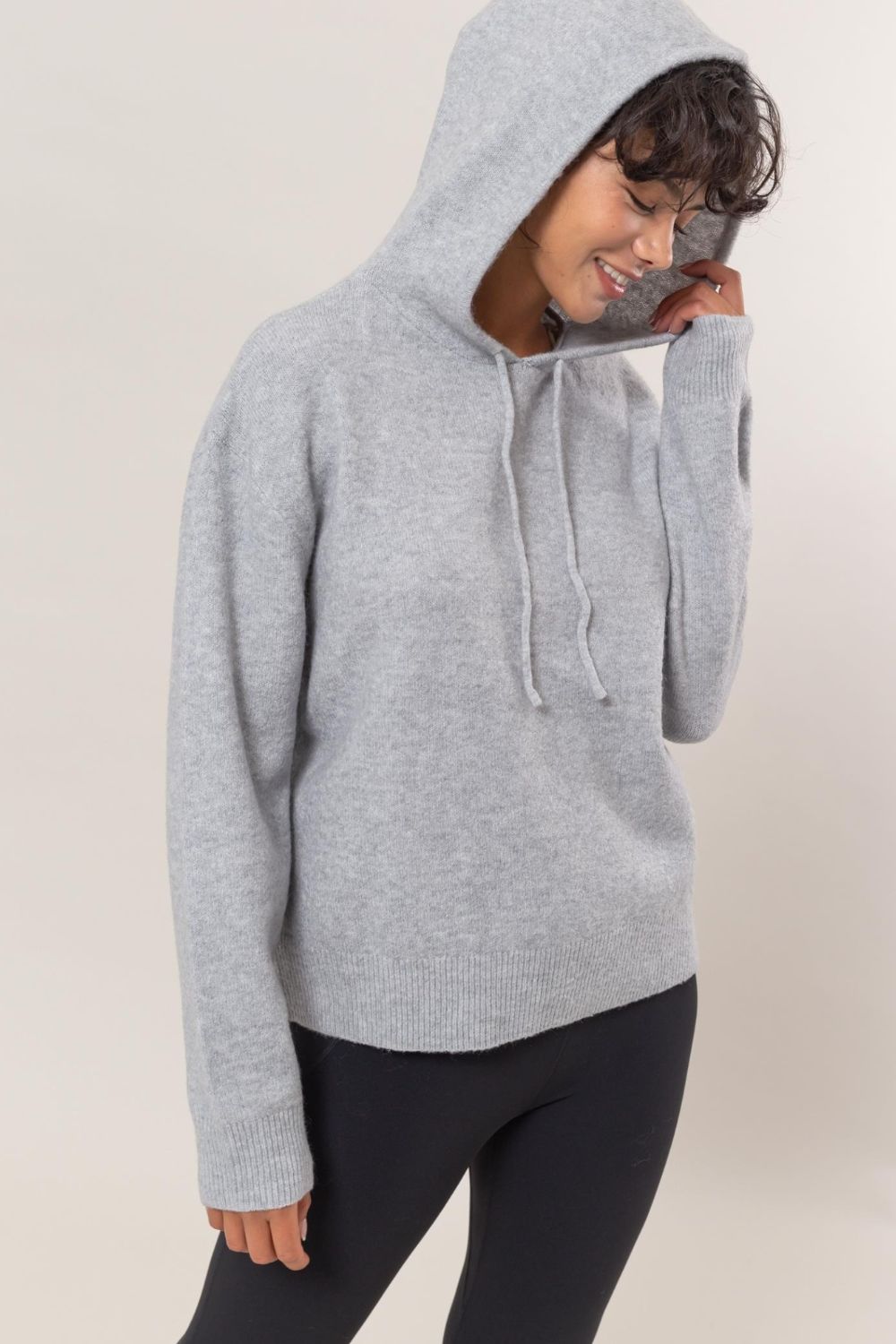 Woman wearing HYFVE drawstring long sleeve hooded sweater in gray, styled with black leggings for a cozy, casual look.