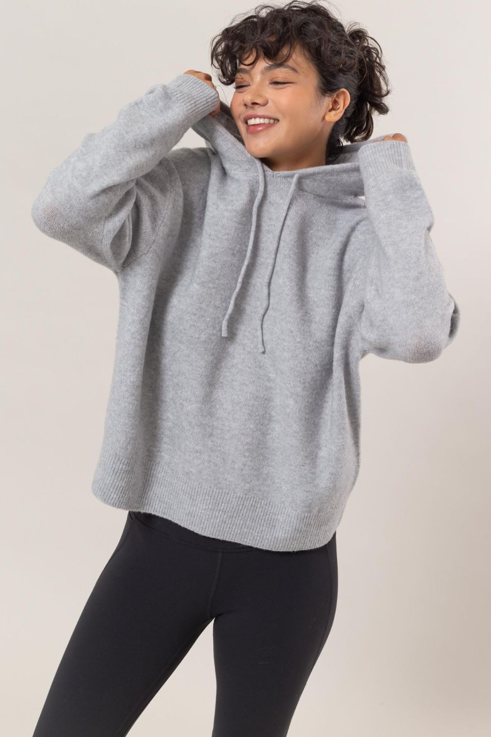 Woman wearing a gray drawstring long sleeve hooded sweater looking cozy and stylish, paired with black leggings.