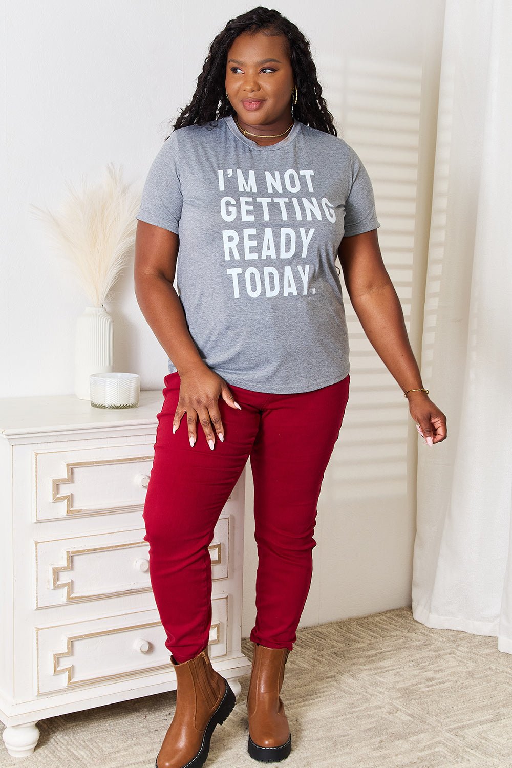 Woman wearing "I'M NOT GETTING READY TODAY" graphic T-shirt, styled with red pants and brown boots, standing in a modern room.