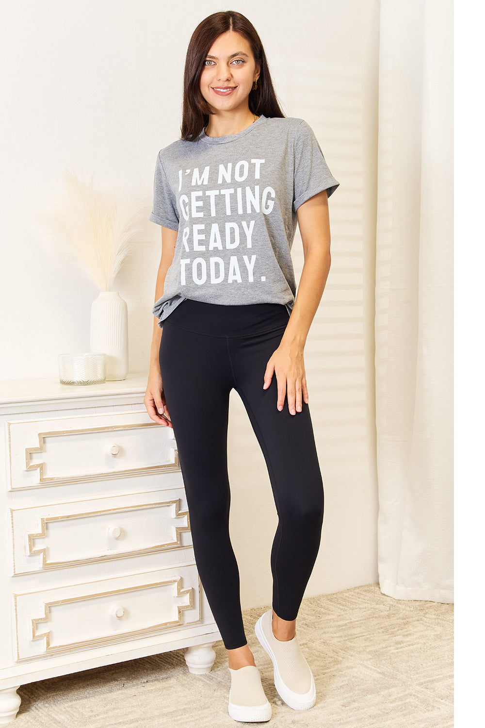 Woman wearing "I'M NOT GETTING READY TODAY" graphic t-shirt with black leggings in a casual home setting.