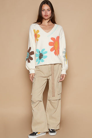Woman wearing an ivory V-neck long sleeve sweater with colorful flower fringe design, paired with beige cargo pants and sneakers.