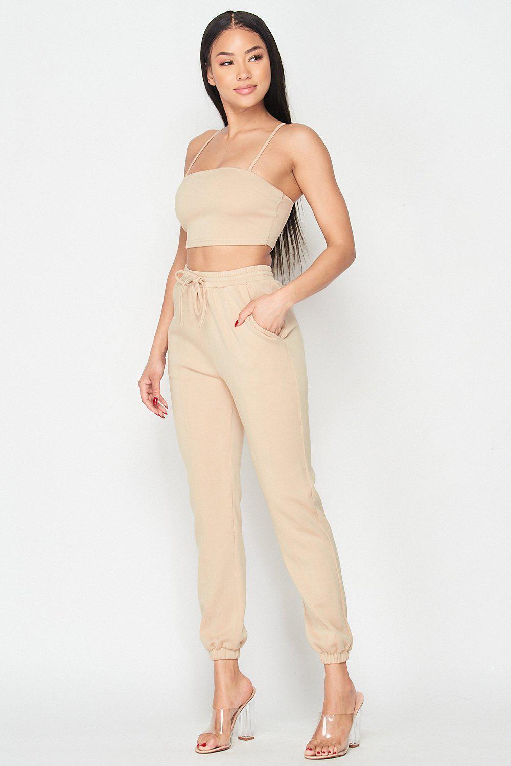 Woman wearing Keep It Cute Two-Piece Set in beige, featuring a spaghetti strap crop top and jogger pants, styled with clear heels.