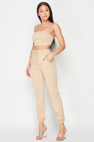 Woman wearing Keep It Cute Two-Piece Set in beige, featuring a spaghetti strap crop top and jogger pants, styled with clear heels.