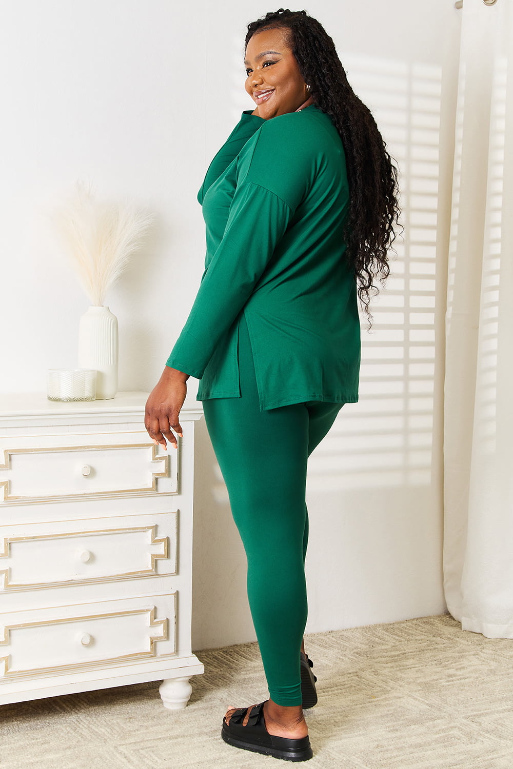 Woman wearing green long sleeve top and leggings set, standing by a white dresser in a sunlit room.