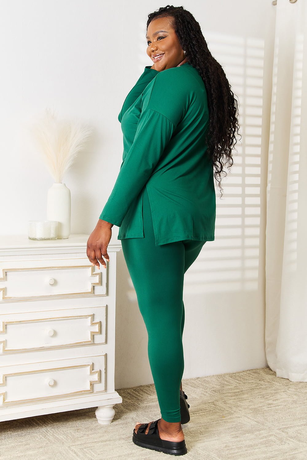 Woman wearing green long sleeve top and leggings set, standing by a white dresser in a sunlit room.