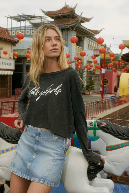 Woman in "Let's Go Girls" metallic graphic tee and denim skirt, standing in vibrant outdoor setting with red lanterns.