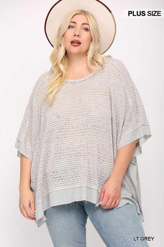 Plus size model wearing light knit woven boxy top with poncho sleeves in light grey, styled with jeans and a hat.