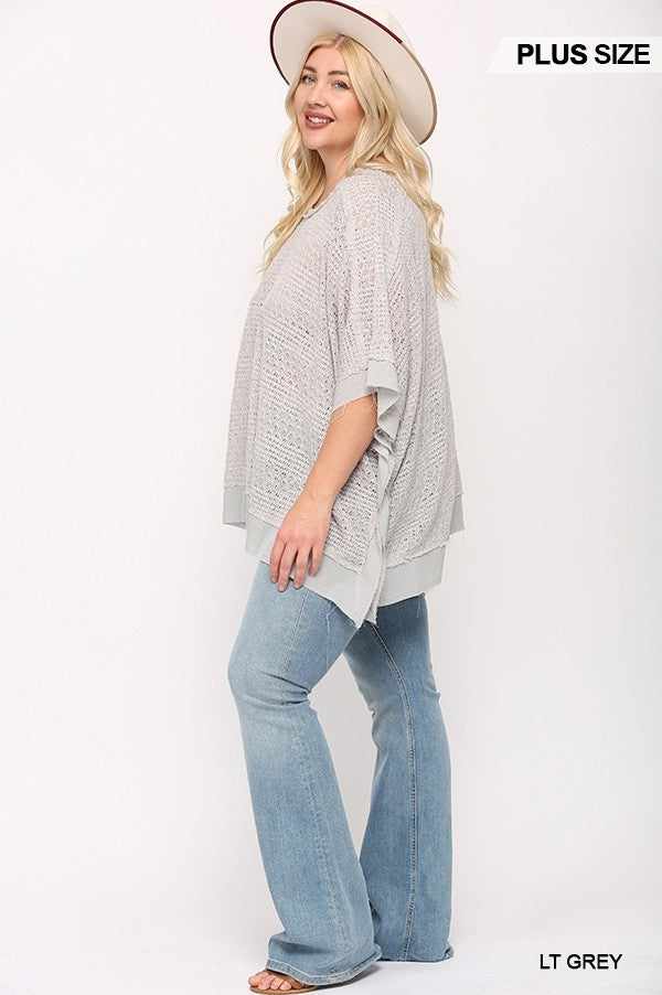 Plus size woman in light grey knit and woven boxy top with poncho sleeves and jeans, wearing a hat.