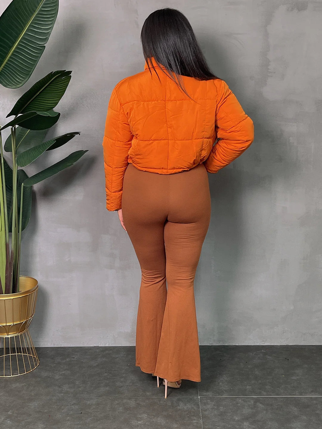 Woman wearing an orange lightweight jacket and brown flared pants, standing in front of a gray wall with green plants.