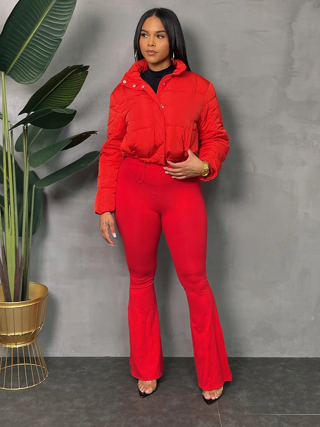 Woman in a vibrant red casual outfit with a lightweight jacket, standing next to a potted plant against a gray background.