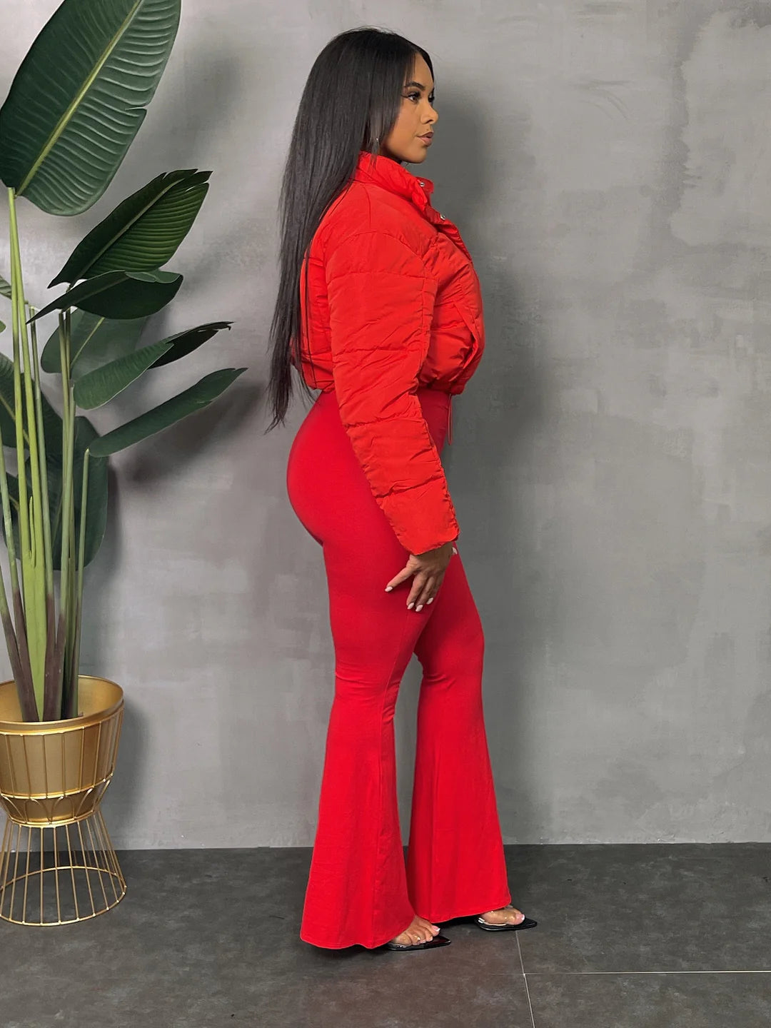 Woman in red flared pants and a puffer jacket standing near potted plant, showcasing chic and vibrant fashion style.