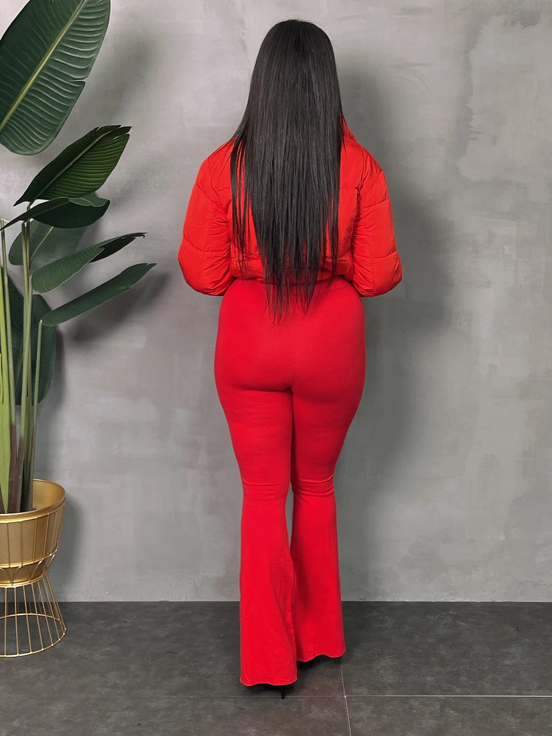 Woman in red lightweight jacket and flared pants stands against a gray background with a potted plant on the side.