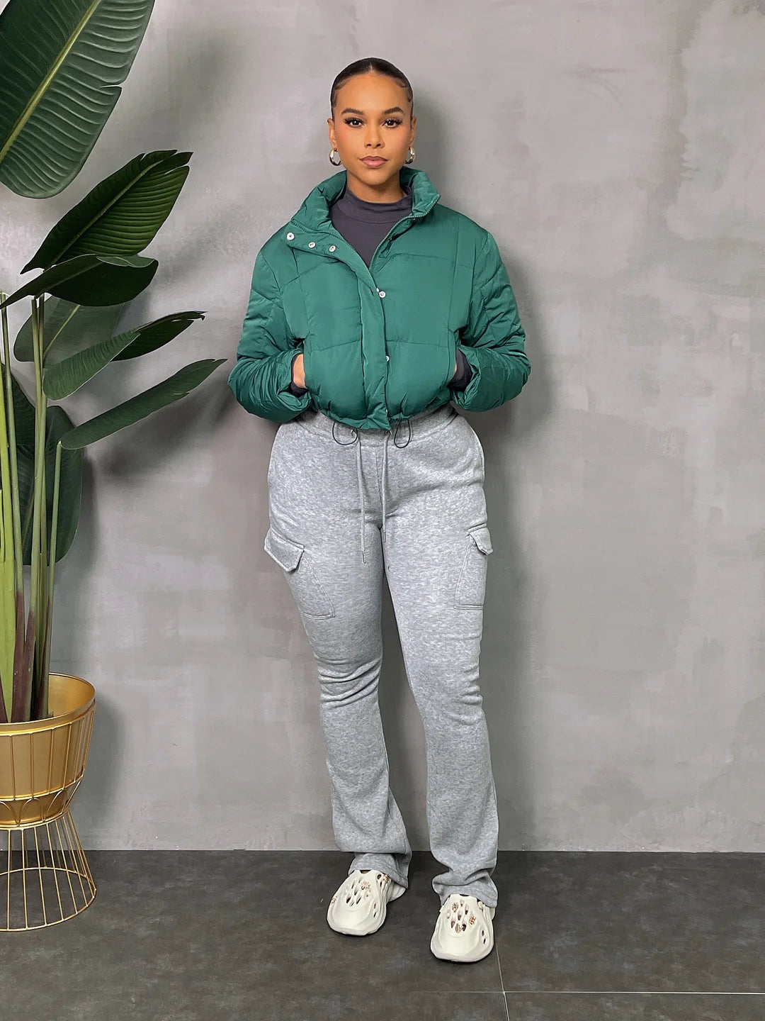 Woman in teal puffer jacket with gray joggers and white sneakers standing indoors near green plant.