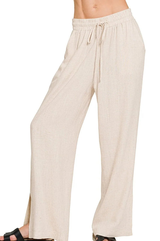 Woman wearing cream-colored wide-leg pants with drawstring waist and side slits, paired with black sandals, standing against white background.