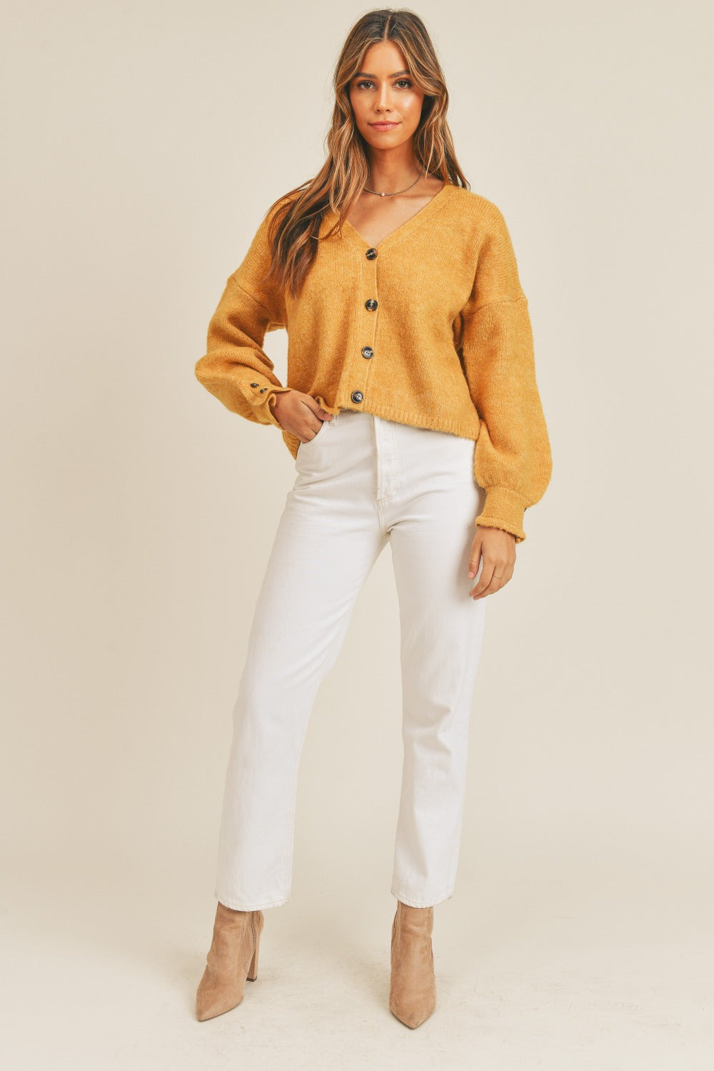 Woman wearing a mustard yellow long sleeve button-down sweater cardigan with white pants and beige boots.