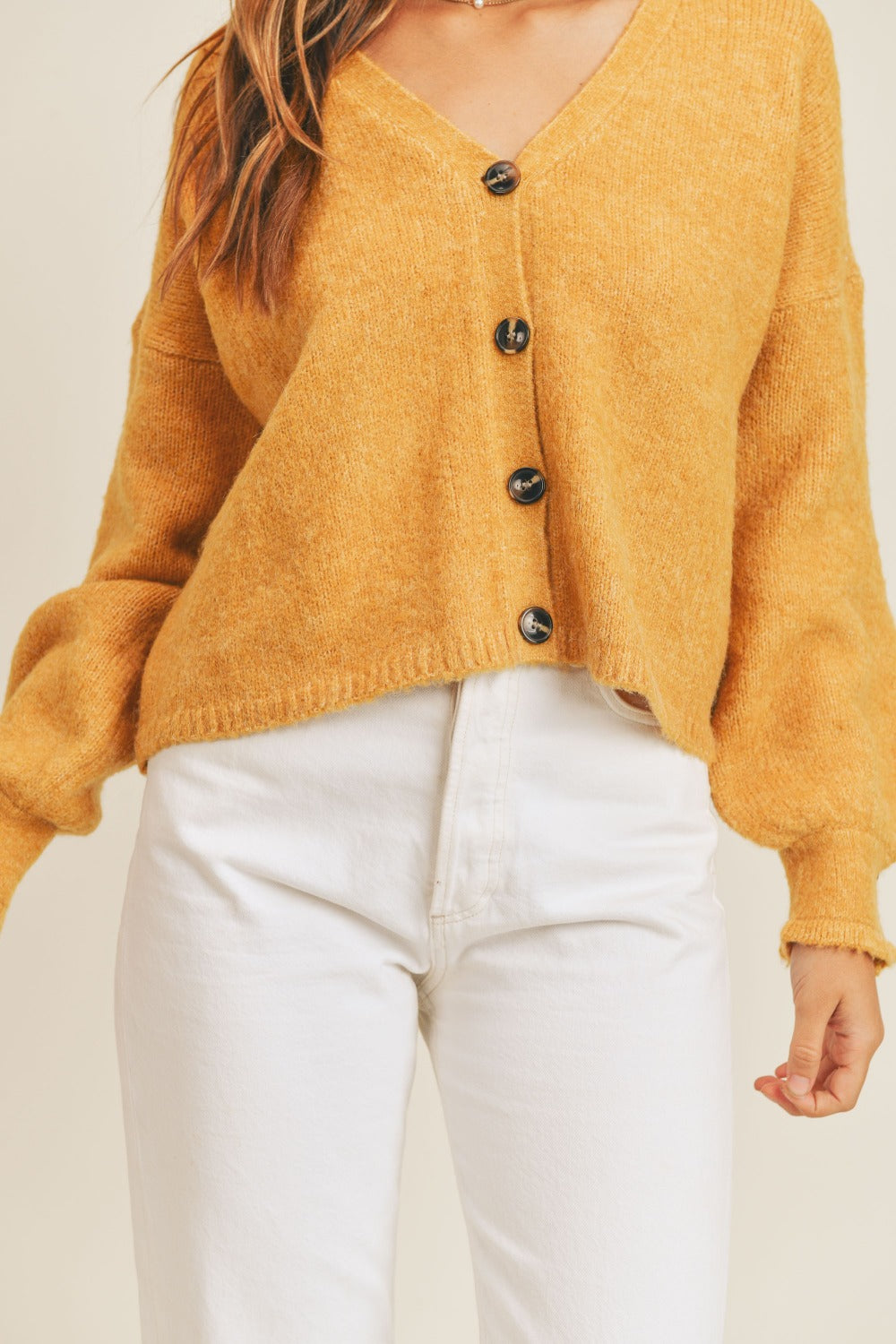 Woman wearing a cozy long sleeve button down sweater cardigan in mustard yellow, paired with white pants.