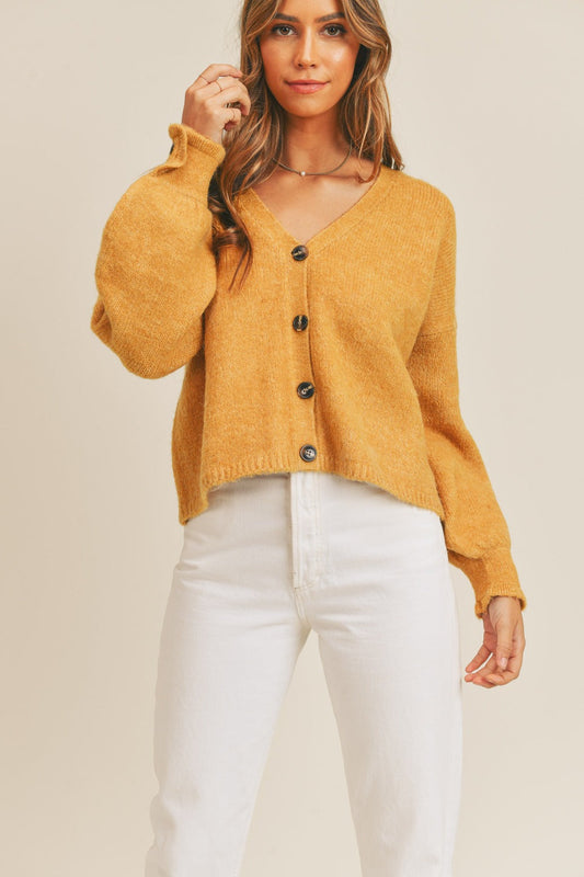 Woman wearing a mustard yellow long sleeve button down sweater cardigan made of acrylic, polyester, and spandex, paired with white pants.