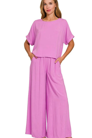 Woman wearing a bright pink two-piece outfit with a loose-fitting short-sleeve top and wide-leg pants, standing with hands in pockets against a whi...