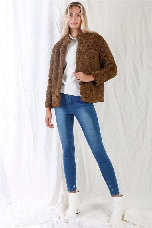 Woman wearing mid blue high-waisted skinny jeans and a brown jacket, styled with white boots, standing against a draped white background.