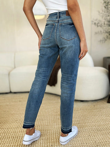 Woman wearing mid rise rigid magic release hem jeans with moderate stretch, showcasing back pockets, in a cozy living room setting.