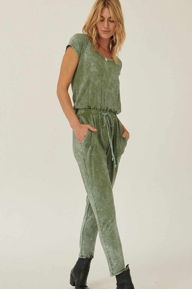 Woman wearing olive mineral washed finish knit jumpsuit with scoop neckline, short sleeves, drawstring waist, and tapered leg cuffs.