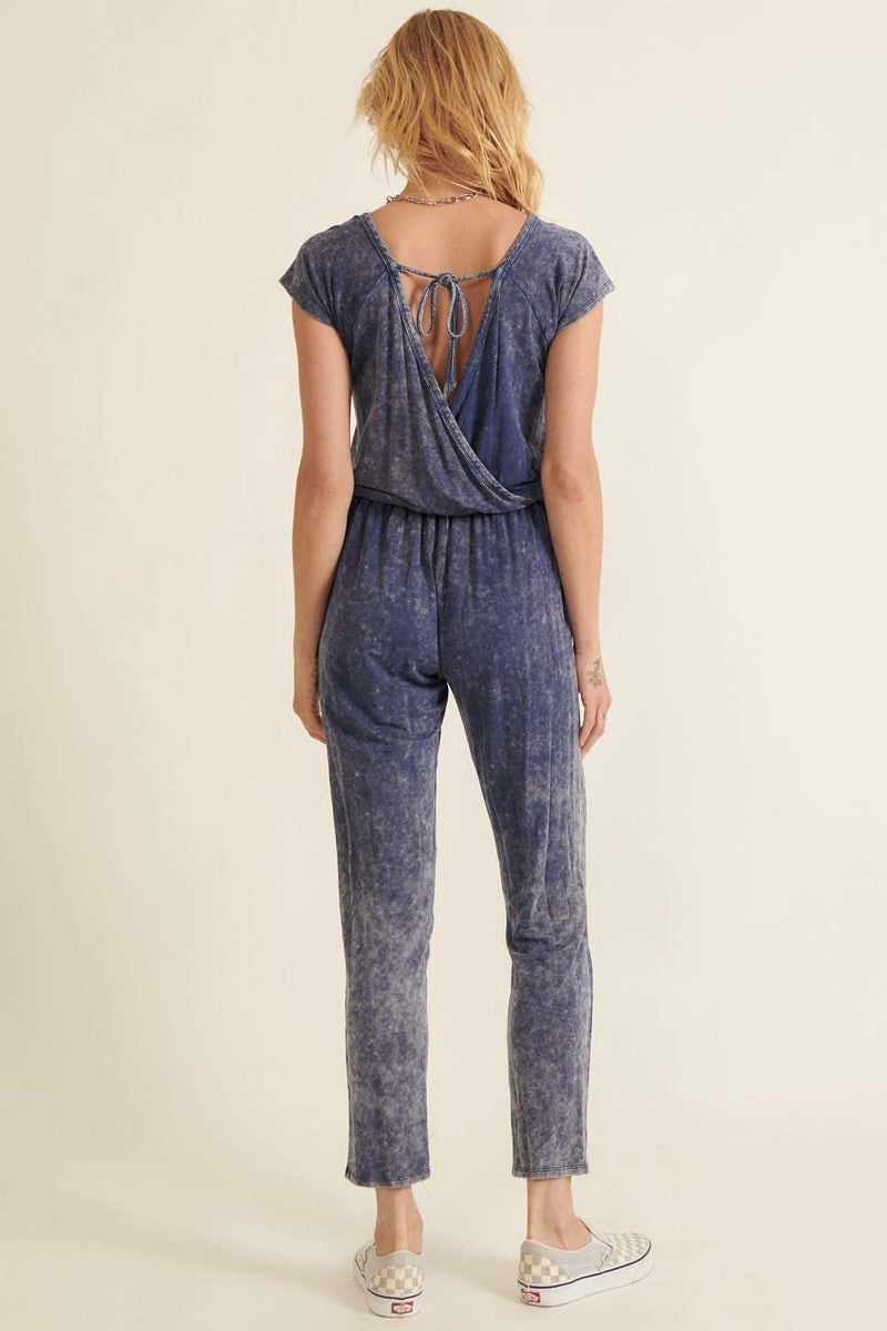 Woman wearing a denim blue mineral washed knit jumpsuit with an open back, drawstring waist, and cuffed legs.