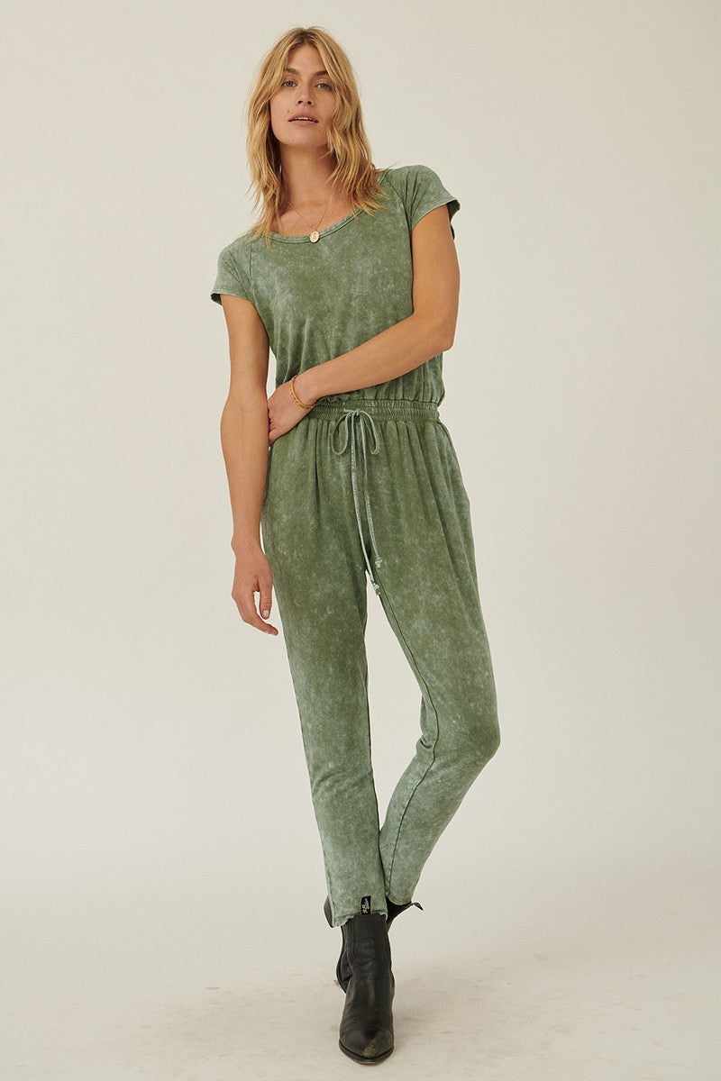 Olive mineral washed knit jumpsuit with scoop neckline, short sleeves, elastic drawstring waist, tapered legs, and open back.