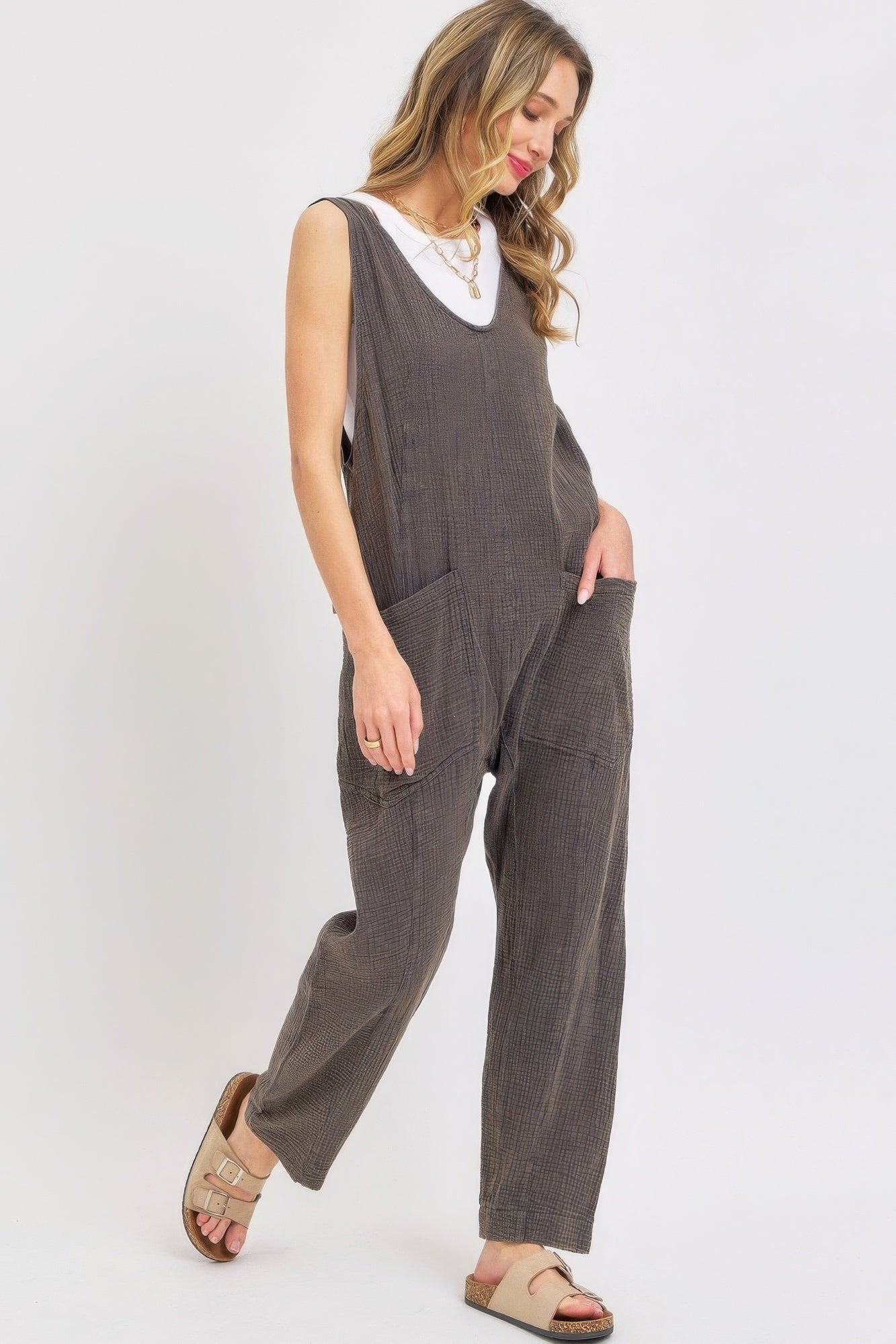 Woman wearing a mineral washed summer jumpsuit with adjustable straps and oversized pockets, styled with sandals and a white top.