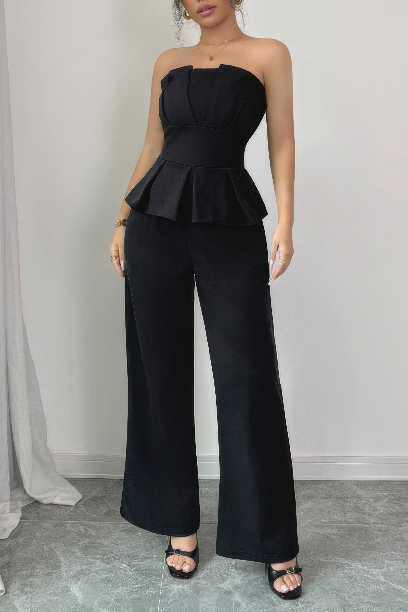 Woman wearing black strapless peplum top with wide-leg pants, standing against white wall on marble flooring, wearing black strappy heels and gold...