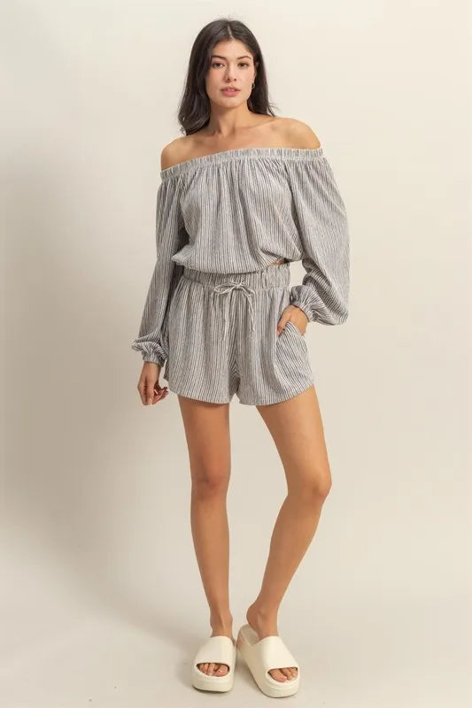 Woman wearing gray striped off-shoulder romper with long sleeves and drawstring waist, paired with white slide sandals, standing against beige...