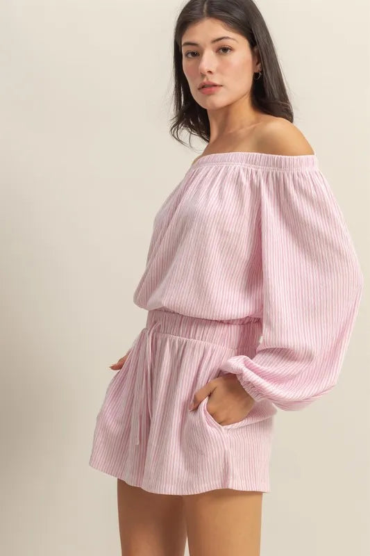 Woman wearing pink and white striped off-shoulder romper with puffy sleeves, posing against beige background.