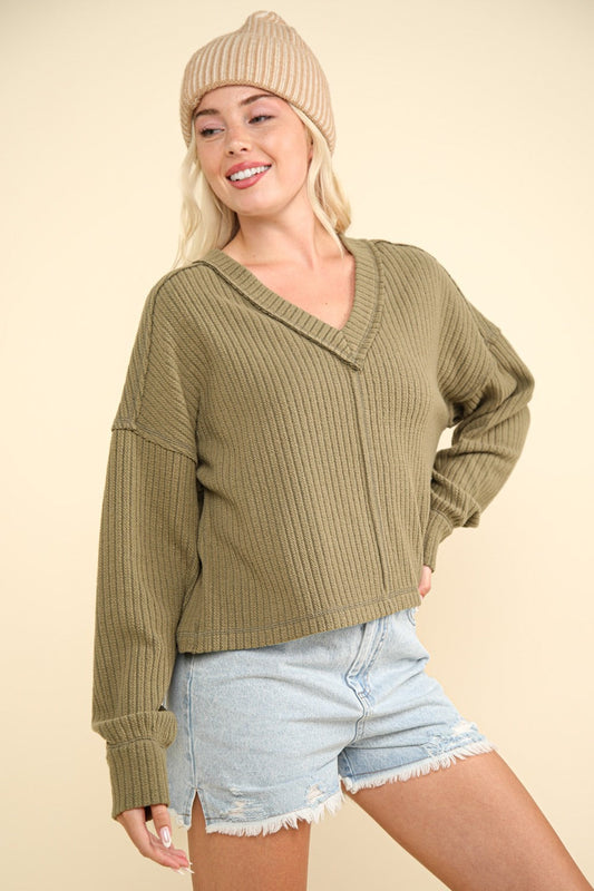 Woman wearing olive ribbed V-neck knit top with exposed seam, paired with denim shorts and a beige beanie.