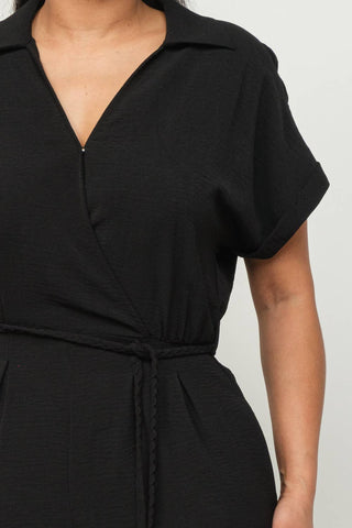 Black wrap top jumpsuit with waist tie and short sleeves, featuring inside button detail and collared neckline.