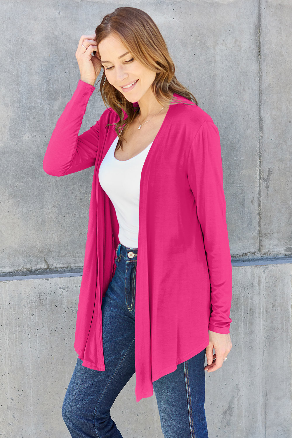 Woman wearing pink open front long sleeve cardigan, paired with white top and jeans, against a gray concrete background.