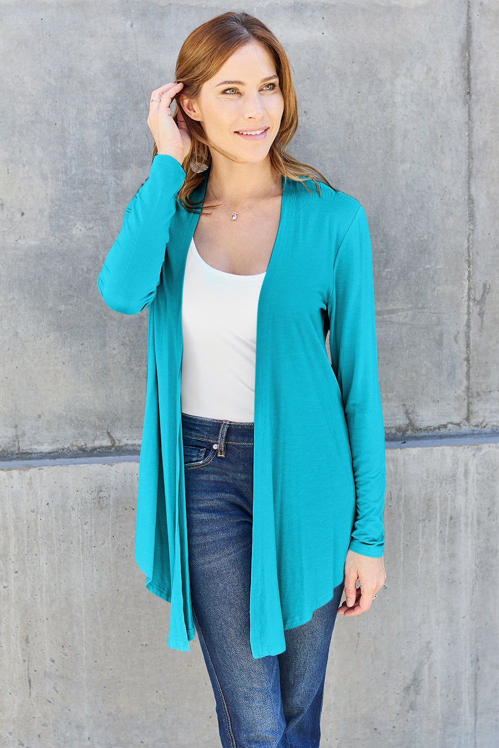 Woman wearing an open front long sleeve cardigan in teal over a white top and jeans against a concrete background