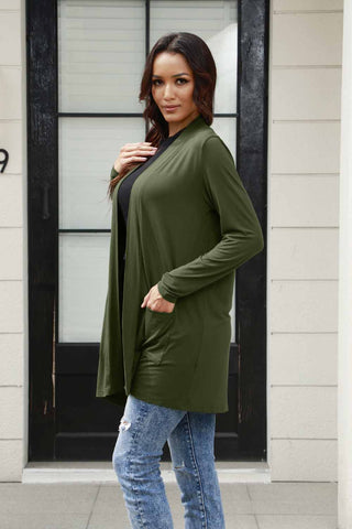 Woman wearing a green open front long sleeve cardigan with pockets, paired with blue jeans, standing in front of a door.