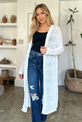 Woman wearing openwork longline cardigan over black top and jeans indoors.