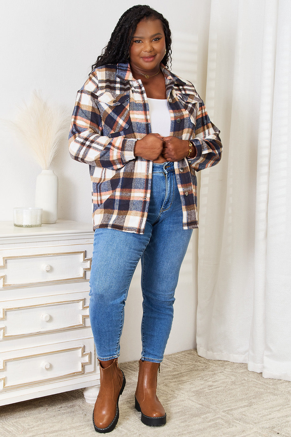 Woman wearing Double Take Plaid Button Front Shirt Jacket with breast pockets, paired with jeans and brown boots in a stylish room.