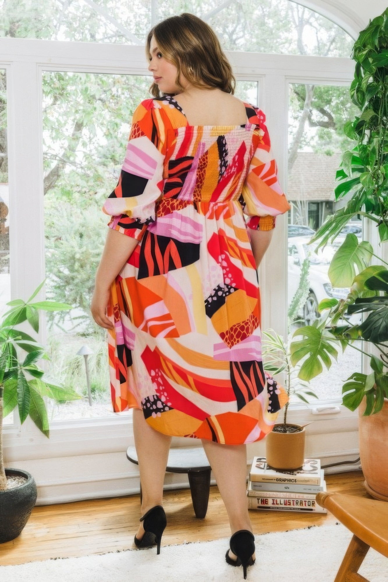 Plus size woman wearing coral floral print midi dress with square neckline and puff sleeves, standing in bright room with plants.