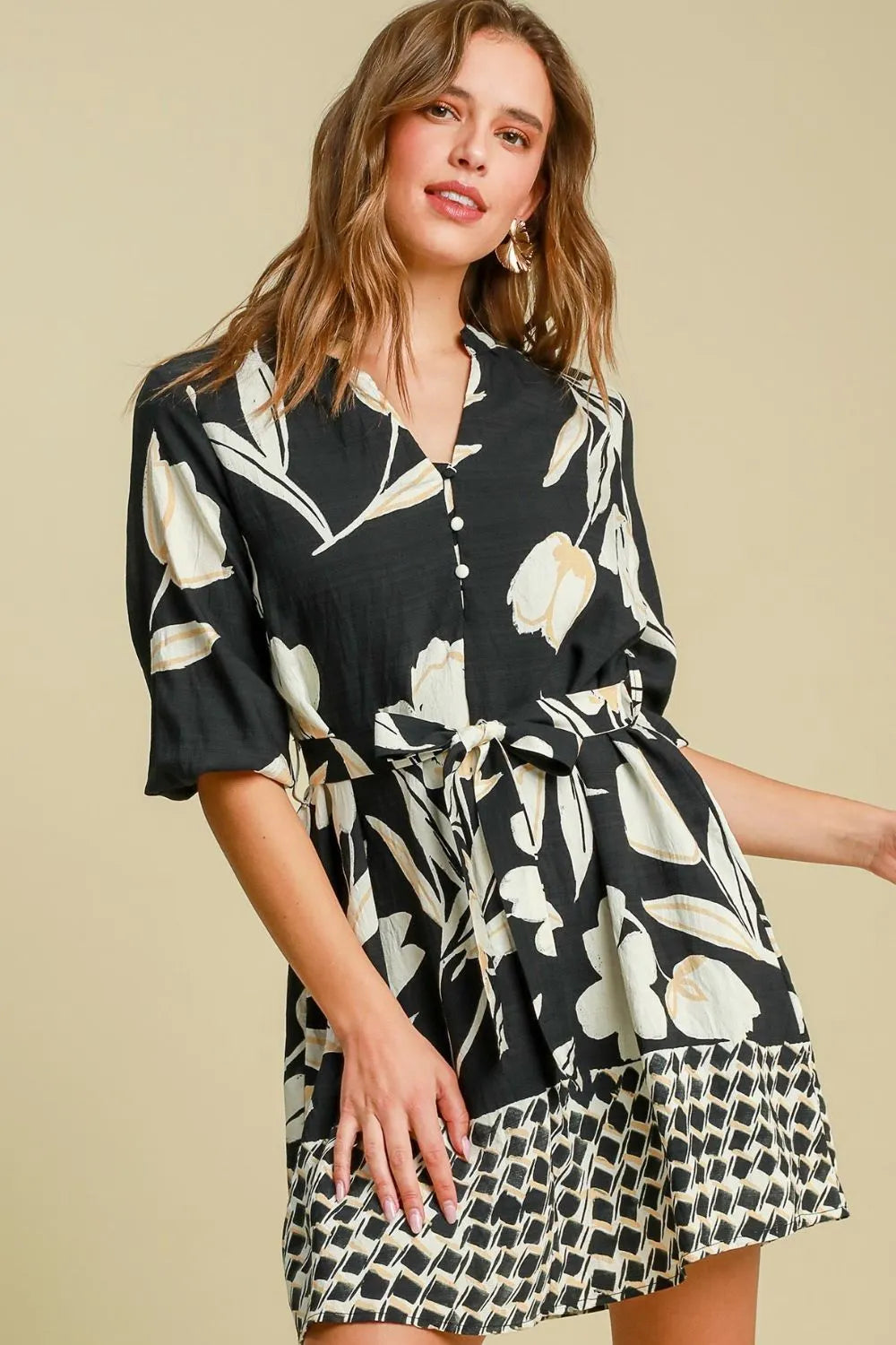 Woman in black floral print dress with white leaf pattern and geometric border hem, three-quarter sleeves, tie waist, standing against beige backgr...