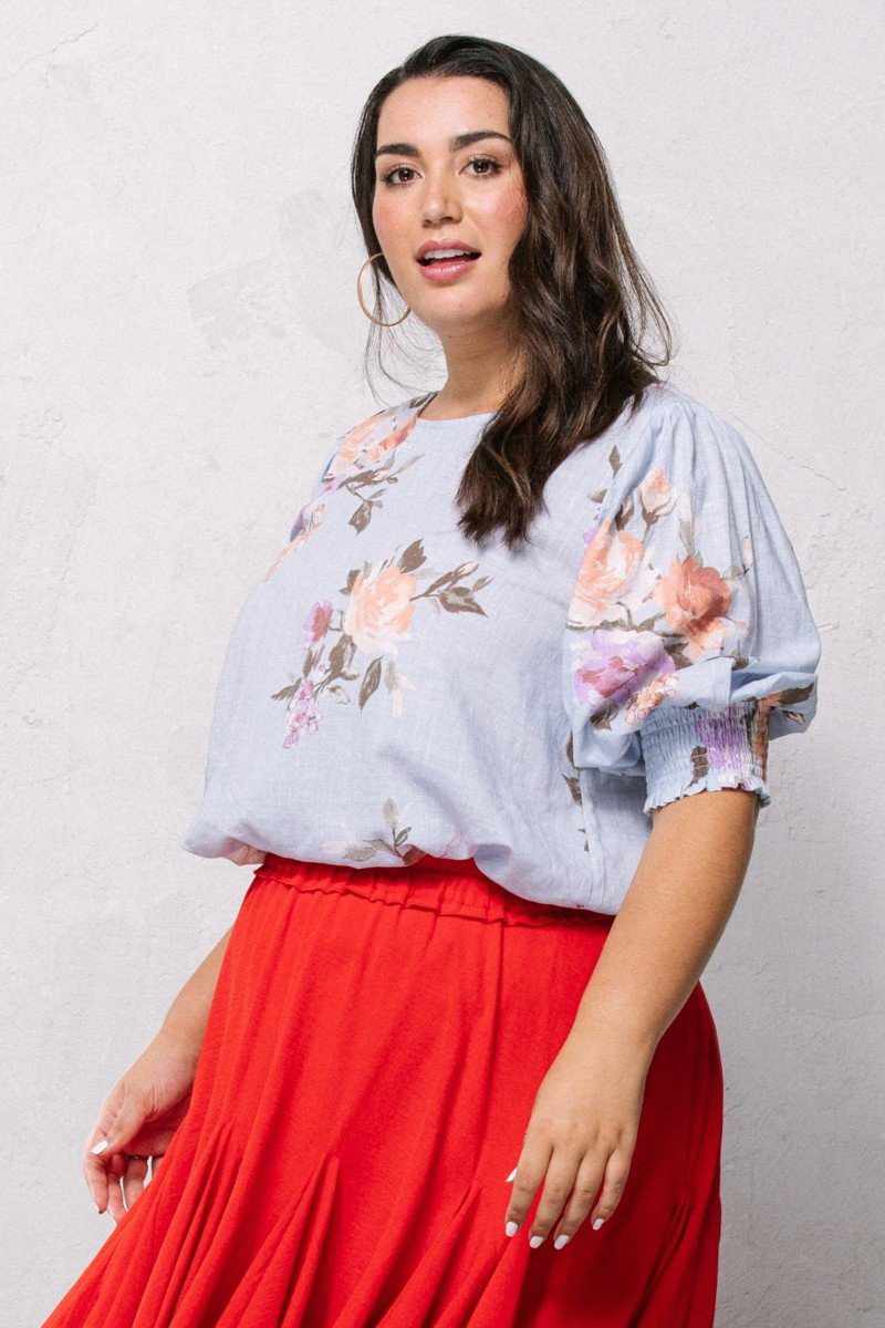 Woman wearing a blue floral printed woven blouse with short puff sleeves and a red skirt, featuring a round neckline and smocked hemline.