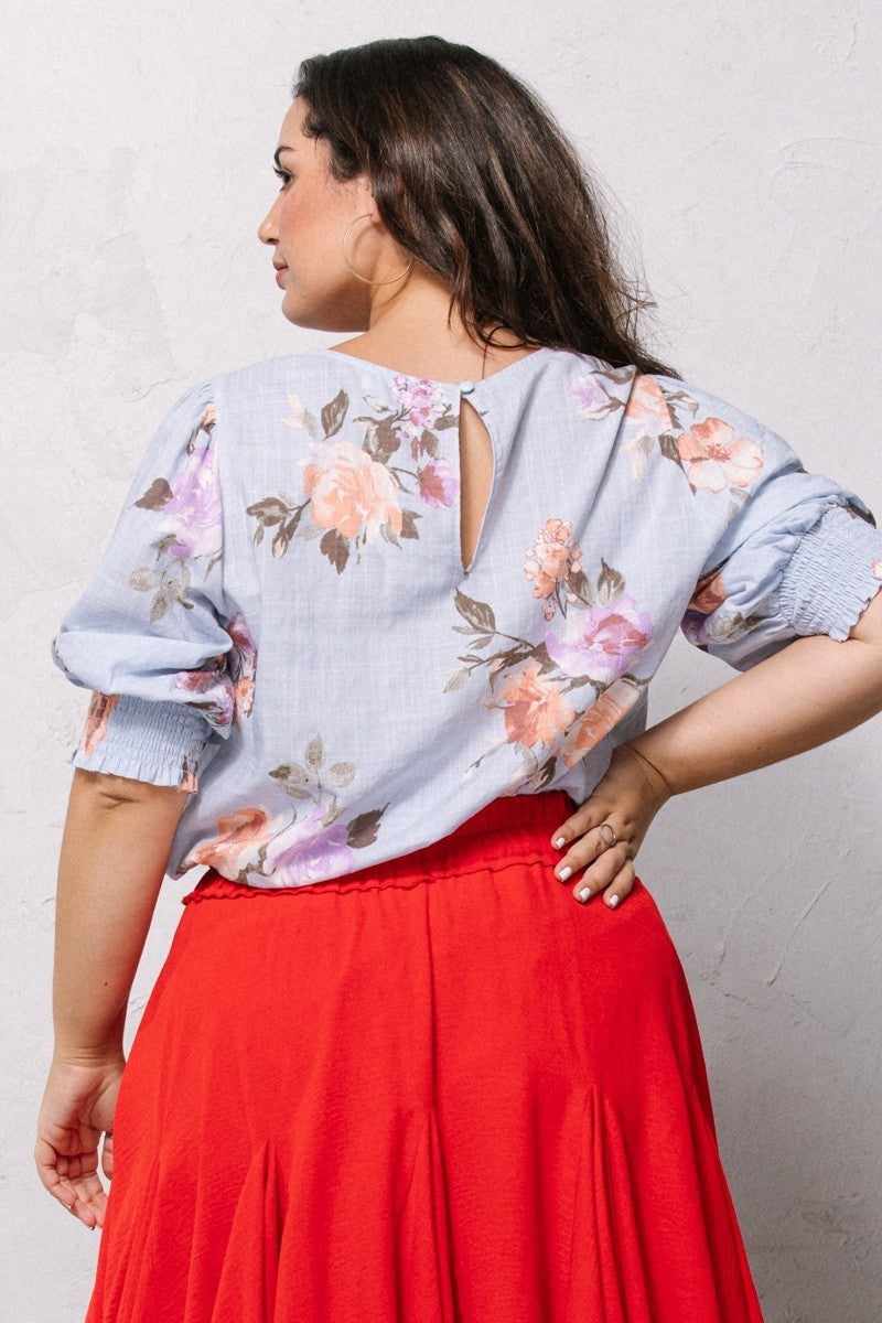 Plus-size woman modeling a blue floral-printed woven blouse with short puff sleeves, back button detail, and smocked hemline.