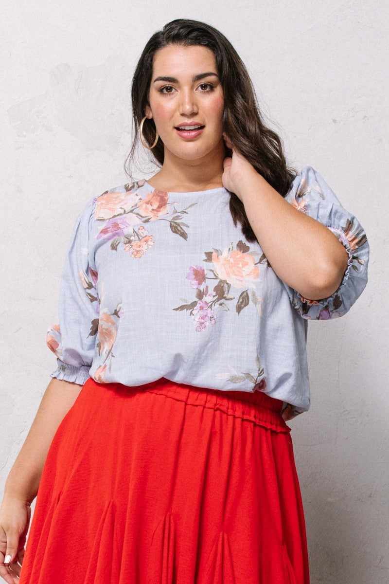 Woman wearing blue floral printed woven blouse with puff sleeves and red skirt, featuring a round neckline and smocked hemline.