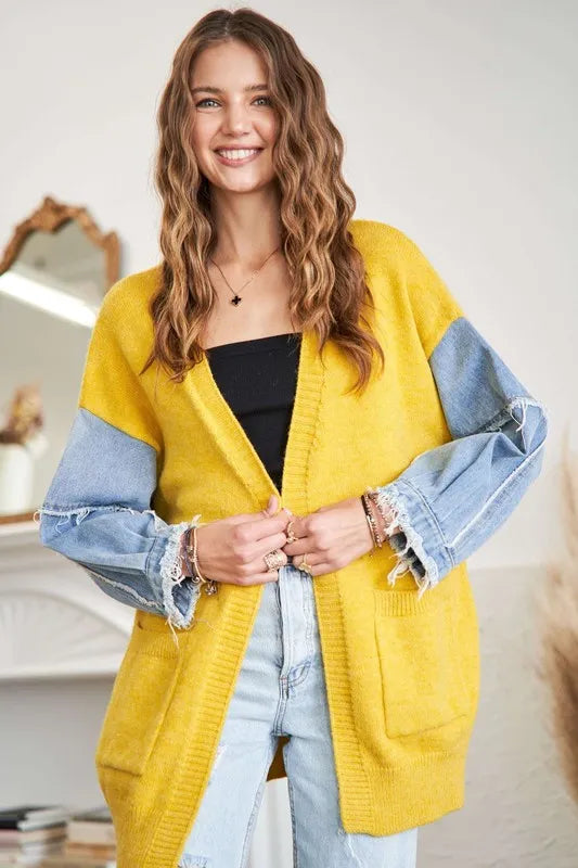 Woman with long wavy brown hair smiling, wearing a bright yellow cardigan over a black top and light blue jeans, standing in a bright indoor settin...
