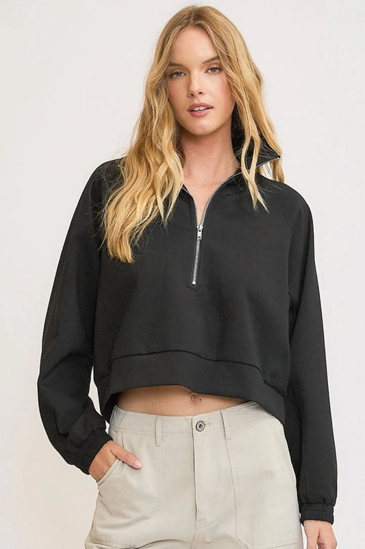 Woman with long blonde hair wearing a black cropped zip-up sweatshirt and beige pants, standing against a light gray background with one hand in po...